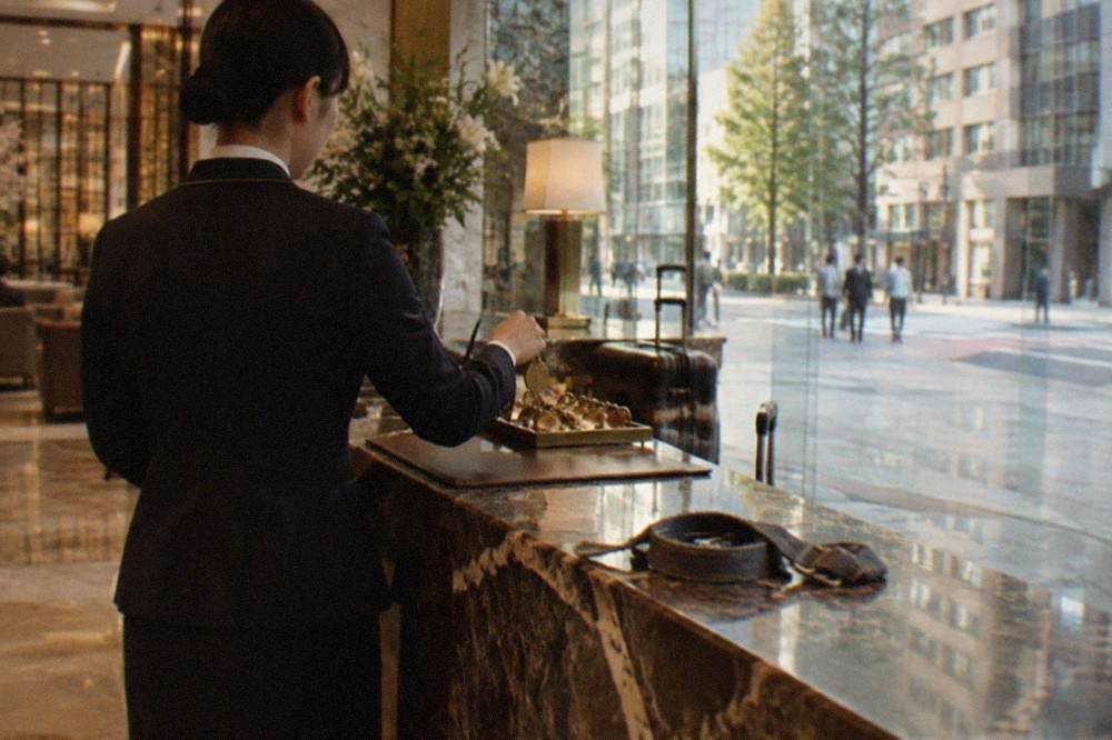 A hotel bell desk attendant taking a numbered luggage tag from a counter beside a suitcase