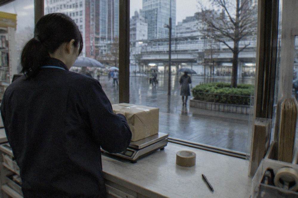 A convenience store counter in Tokyo with a shipping clerk weighing a customer's parcel on a small scale