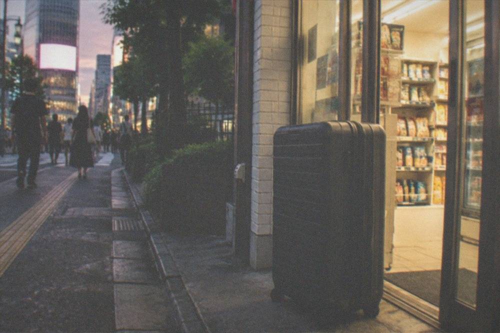 A large hard-shell suitcase standing next to a small convenience store entrance in a Tokyo street