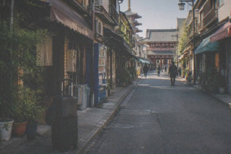 A wheeled suitcase and a smaller shoulder bag set down on the ground at a quiet Tokyo backstreet of low shopfronts, one of the visitor districts where bags can be left for the day.