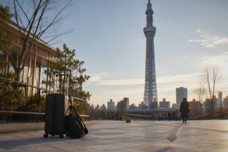 A wheeled suitcase and a smaller shoulder bag set down on the ground at a paved plaza in Tokyo with a tall broadcasting tower rising in the distance, standing for the drop-off points near the city landmarks.