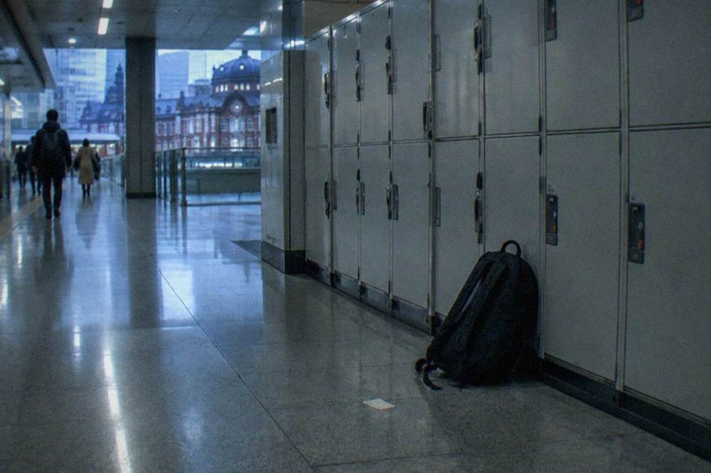A bank of grey coin lockers in a Tokyo subway corridor with a traveller resting a backpack against one.