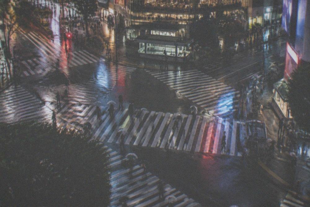 A wide Tokyo intersection at night seen from above with illuminated crossings and scattered pedestrians.