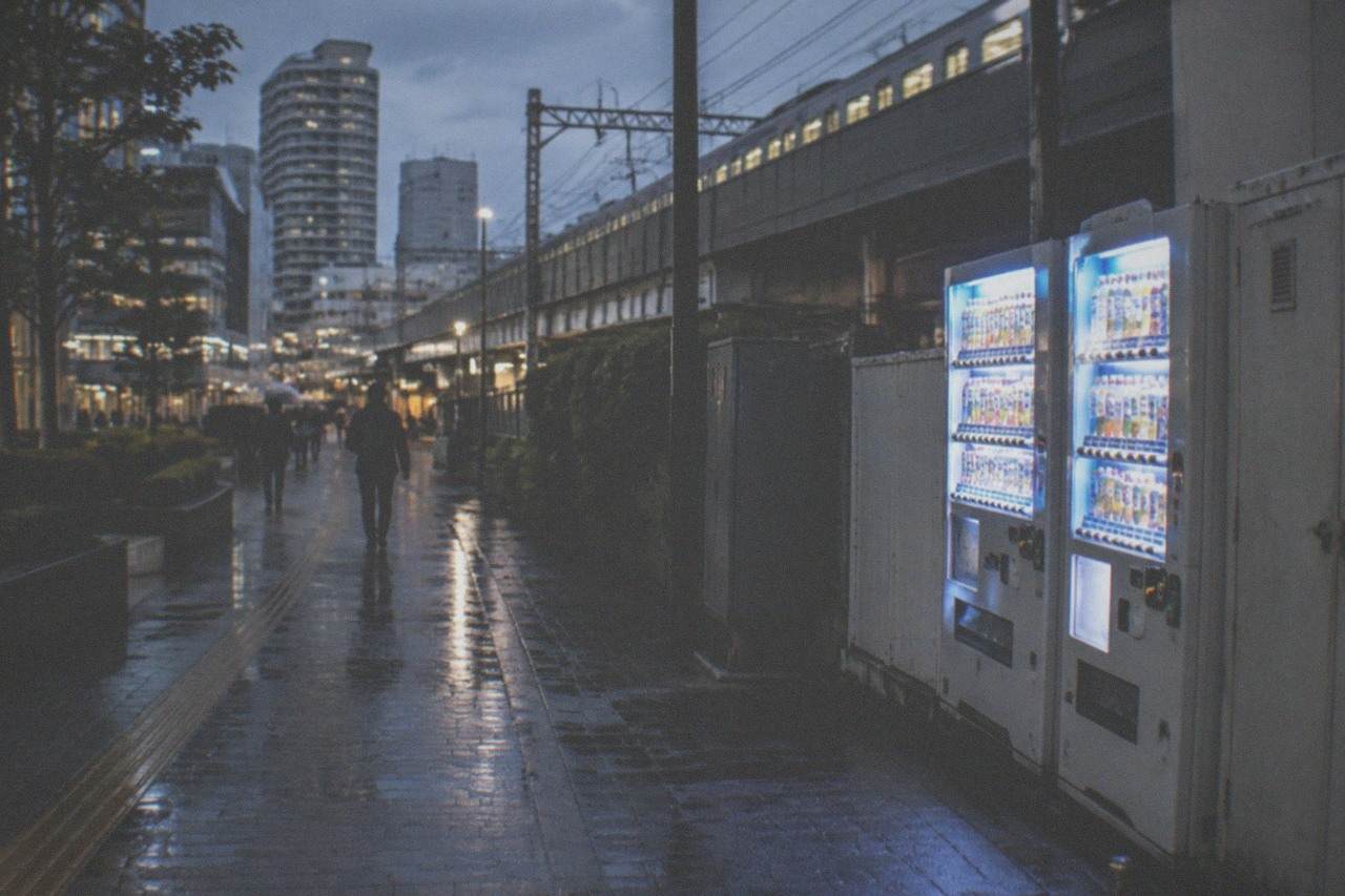 A quiet Tokyo street at dusk with vending machines glowing under railway overhead lines.