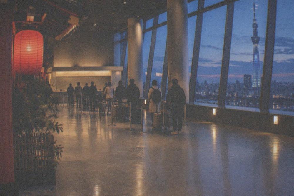 An airport terminal hall at dusk with an airline check-in counter and travellers queuing with trolleys