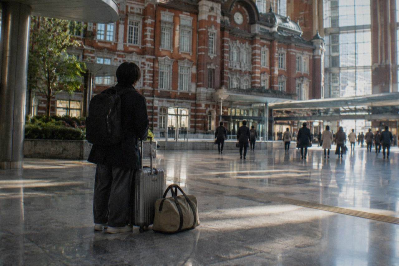 A wheeled suitcase and a duffel bag standing on the tiled floor of a Tokyo railway station concourse beside a traveller seen from behind, the gap between arriving and hotel check-in that luggage storage covers.